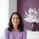 Portrait of a smiling woman with light brown skin and dark, wavy hair, wearing a soft lavender top against a bright white and